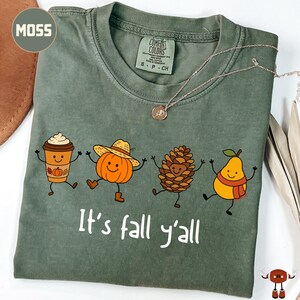 May include: A moss green t-shirt featuring a graphic of a coffee cup, a pumpkin wearing a straw hat, a pinecone, and a pear wearing a scarf, all with smiling faces. The text "It's fall y'all" is printed in white below the characters.