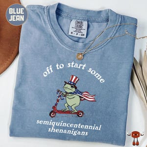 Könnte beinhalten: Blaues Jeans-T-Shirt mit einer Grafik einer Froschfigur auf einem roten Roller, die einen patriotischen Hut und Umhang trägt. Der Text lautet "off to start some semiquincentennial shenanigans". Eine goldene Halskette liegt auf dem Shirt.