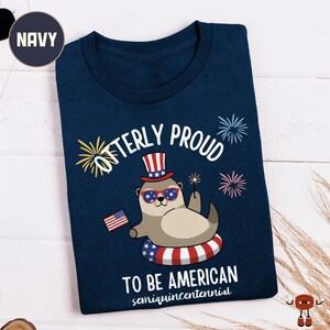 May include: Navy blue t-shirt featuring a cartoon otter wearing sunglasses, a patriotic hat, and an inner tube. The text reads "Otterly Proud To Be American" with fireworks and an American flag graphic. The shirt is folded.