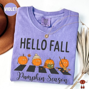 May include: A violet t-shirt featuring the text HELLO FALL and Pumpkin Season in black. The graphic shows four orange pumpkins walking across a black zebra crossing. One pumpkin wears a beanie, one holds a leaf, one reads a book, and one holds a broom.