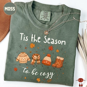 May include: A moss green t-shirt featuring the white text 'Tis the Season to be cozy' and a graphic of a smiling sweater, scarf, beanie, and boots surrounded by falling autumn leaves. The shirt has a crew neckline and a Comfort Colors brand label.