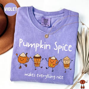 May include: A violet short-sleeve t-shirt featuring the text "Pumpkin Spice makes everything nice" and a graphic of four animated coffee cups with faces. The shirt has a Comfort Colors brand label at the neck and a thin gold-toned necklace.