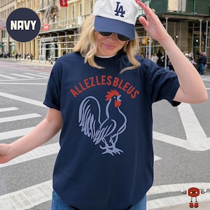 May include: A navy blue short-sleeve t-shirt featuring a graphic of a rooster in light blue with a red comb and wattle. The text "ALLEZLESBLEUS" is printed in red in an arc above the rooster graphic. The shirt has a crew neckline and a relaxed fit.