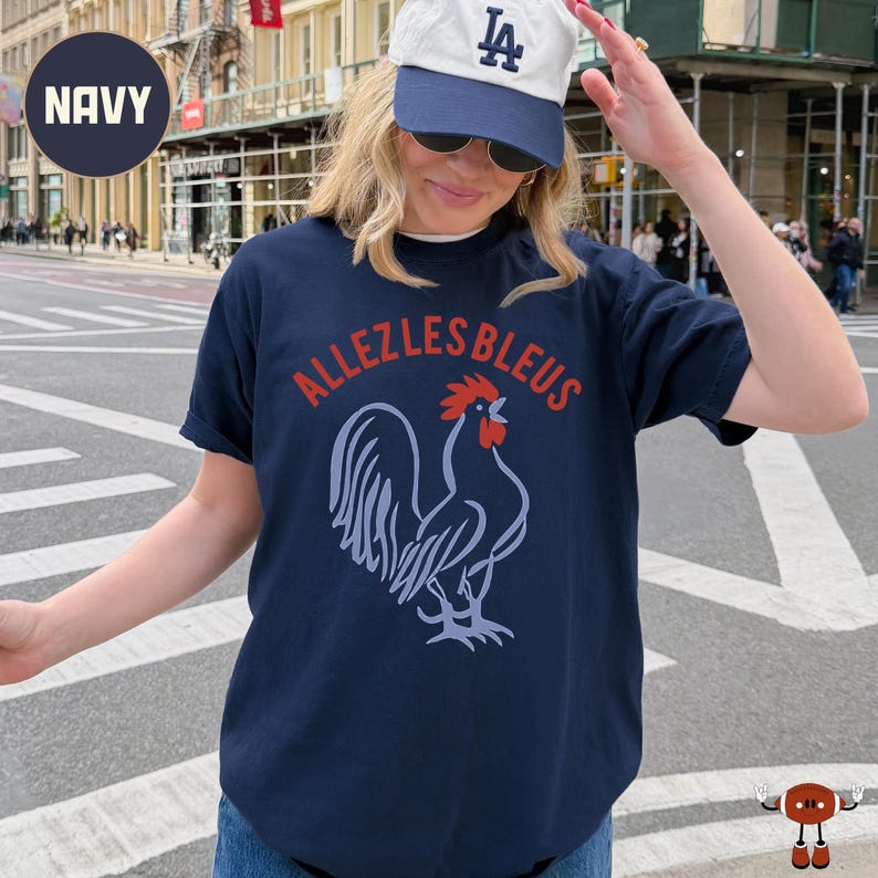 May include: A navy blue short-sleeve t-shirt featuring a graphic of a rooster in light blue with a red comb and wattle. The text "ALLEZLESBLEUS" is printed in red in an arc above the rooster graphic. The shirt has a crew neckline and a relaxed fit.
