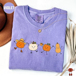 May include: A violet short-sleeved t-shirt featuring a graphic of four cartoon pumpkins with arms and legs. The first pumpkin wears a straw hat and boots, the second is white, the third holds a mug, and the fourth is a tall butternut squash.