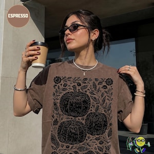 May include: A brown short-sleeved t-shirt featuring a black graphic print of three pumpkins surrounded by floral and leaf patterns. The shirt has a crew neckline and a relaxed fit. The word ESPRESSO is printed in the upper left corner of the image.