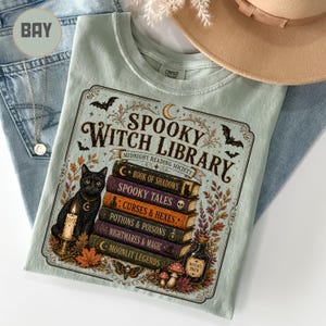 May include: A light green t-shirt featuring a graphic print with the text SPOOKY WITCH LIBRARY and MIDNIGHT READING SOCIETY. The design includes a stack of books titled Book of Shadows, Spooky Tales, Curses & Hexes, Potions & Poisons, Nightmares & Magic, and Moonlit Legends, alongside a black cat, a candle, a bottle labeled Witch's Brew, bats, and autumn leaves.