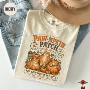 May include: An ivory t-shirt featuring a graphic print with the text PAW-MPKIN PATCH. The design includes three orange tabby cats sitting among pumpkins, with additional text reading LOCALLY GROWN, PICK OF THE PATCH, and THE CHONKIEST IN THE PATCH.