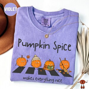 May include: A violet short-sleeve t-shirt with a graphic print of four pumpkins walking on a zebra crossing. The text reads "Pumpkin Spice" and "makes everything nice". The shirt features a Comfort Colors brand label at the inner neckline.
