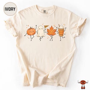 May include: An ivory t-shirt featuring a graphic print of four animated autumn characters: an orange pumpkin, a white ghost holding a pennant that says SPOOKY, an orange maple leaf, and a brown coffee cup, all with smiling faces and thin limbs.