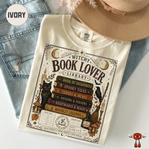 May include: An ivory t-shirt featuring a graphic print titled Witchy Book Lover Library. The design includes a stack of books labeled Book of Shadows, Spooky Tales, Curses & Hexes, Potions & Poisons, Nightmares & Magic, and Moonlit Legends, with a black cat, a raven, a broom, candles, and a library checkout card.