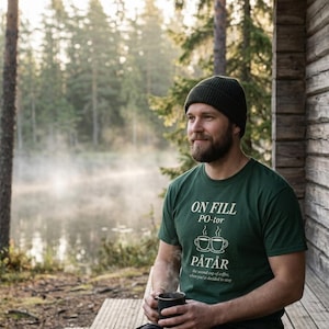 May include: A green t-shirt with the text "ON FILL PO-tor PÅTÅR" and two coffee cup illustrations. The shirt is worn with a dark green beanie. The person is holding a black mug, likely filled with coffee, in a natural outdoor setting.