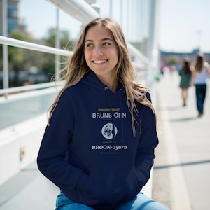 Puede incluir: Una sudadera con capucha azul marino con el texto "BROWN BEAR BRUNBÖFN BROON-ɔyurn" y una ilustración de oso. La sudadera tiene una capucha con cordón y un bolsillo delantero.