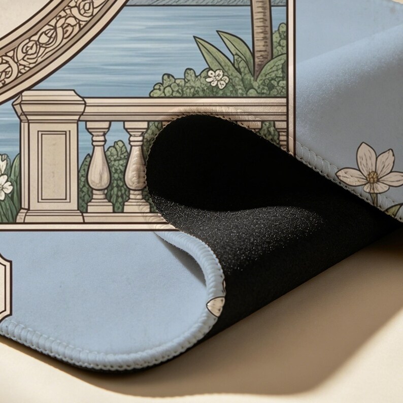 May include: A rectangular desk mat with a light blue surface featuring a printed illustration of a stone balustrade overlooking a body of water with tropical foliage and white flowers. The mat has a thick black foam underside and stitched edges.