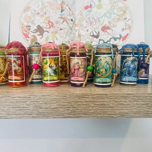 May include: A collection of small glass bottles, each featuring a label with a zodiac sign illustration and name, including Aries, Taurus, Libra, Pisces, Aquarius, and Capricorn. The bottles are sealed with coloured wax and wrapped in twine.