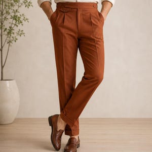 May include: Rust-colored trousers featuring a high-waisted design with a wide waistband, side adjusters, and double pleats at the front. These tailored pants have a straight-leg cut with cuffed hems and are paired with brown leather loafers.
