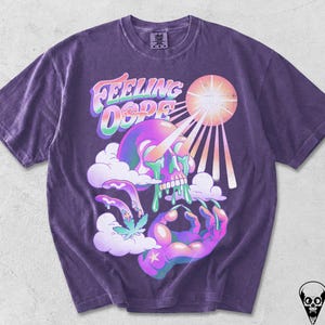 Könnte beinhalten: Ein violettes Kurzarm-T-Shirt mit einem Grafikdruck eines Schädels, aus dessen Augen grüne Flüssigkeit tropft, einer Hand, die eine Okay-Geste macht, Wolken und einer Sonne. Der Text "FEELING DOPE" ist in einer bunten Schrift über dem Schädel gedruckt.