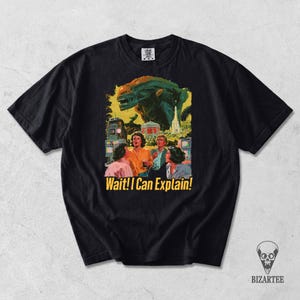Könnte beinhalten: Ein schwarzes Kurzarm-T-Shirt mit einem Grafikdruck, der eine große außerirdische Kreatur zeigt, die über vier Personen in einer Retro-Umgebung thront. Der Text "Wait! I Can Explain!" ist unter der Illustration in Gelb aufgedruckt.