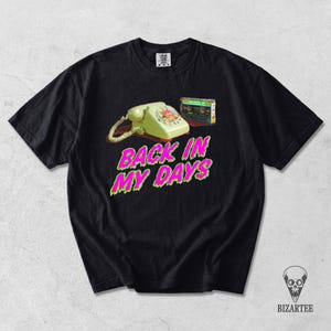 May include: A black short-sleeved t-shirt featuring a graphic of a yellow rotary telephone and a cassette tape. Below the objects, the text "BACK IN MY DAYS" is printed in bright pink with a green outline. The shirt has a crew neck and short sleeves.