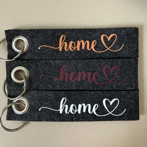 Könnte beinhalten: Drei dunkelgraue Filz-Schlüsselanhänger mit silbernen Metallringen. Jeder Schlüsselanhänger hat das Wort "home" in einer anderen Farbe: Orange, Burgunderrot und Weiß, mit einem Herzsymbol. Die Schlüsselanhänger sind für die Heimdekoration bestimmt.