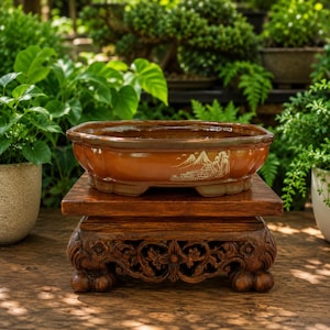 May include: An oval-shaped brown ceramic bonsai pot with a scalloped rim and a white line drawing of a mountain landscape on the side. The pot rests on a dark brown wooden display stand featuring intricate floral carvings and four rounded feet.
