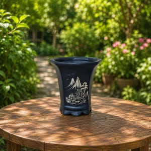 May include: A dark blue ceramic planter with a flared rim and a scalloped base. The front features a white line art illustration of a mountain landscape, including a small building, trees, and birds, rendered in a traditional ink wash style.