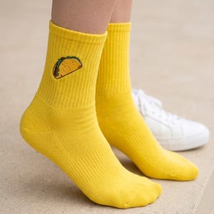 May include: A pair of bright yellow crew socks featuring a small embroidered taco graphic on the outer side of each ankle. The socks have a ribbed texture extending from the cuff down to the mid-foot area, providing a consistent solid color.
