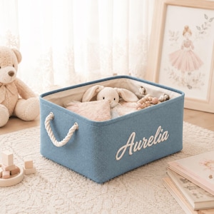Op de afbeelding: Een rechthoekige blauwe stoffen opbergmand met witte touwhandvatten en de naam Aurelia in wit cursief schrift op de voorzijde geborduurd. De mand is gevuld met knuffels en heeft een lichtgekleurde stoffen voering aan de binnenzijde.