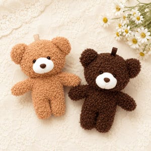 Può includere: Due orsacchiotti di peluche in tessuto riccio e testurizzato. Un orso è marrone chiaro e l'altro è marrone scuro. Entrambi presentano un muso bianco, un piccolo naso marrone scuro e occhi neri. Ogni orso ha un piccolo passante in tessuto.