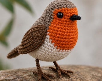 May include: A crochet robin bird figurine featuring a brown head and wings, a bright orange chest, and a cream-colored belly. The bird has a small black beak and round black eyes, with thin brown crocheted legs and feet standing upright.