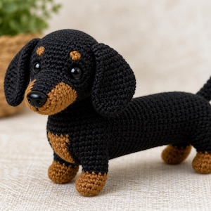 Op de afbeelding: Een gehaakte teckel in zwart en bruin. Het handgemaakte knuffeldier heeft een lang lichaam, korte poten en floppy oren. De hond staat op een neutrale ondergrond, met een vage achtergrond.