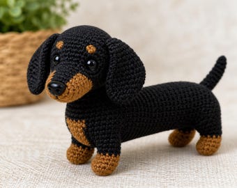 May include: A crocheted dachshund figurine featuring a black body with tan accents on the paws, chest, and above the eyes. The toy has long floppy ears, a small black nose, and dark plastic eyes, standing in a fixed position with a short tail.