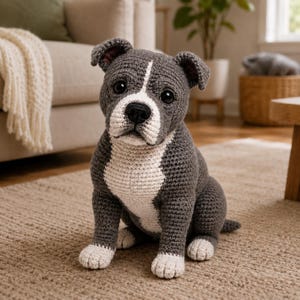 May include: A crocheted dog toy, predominantly grey with white patches on the chest, face, and paws. The dog has black eyes and a black nose. It is sitting on a beige rug. A charming handmade item.