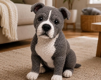 May include: A handmade crochet stuffed animal in the shape of a dog. The dog has dark gray yarn for the body and ears, with a white patch extending from the chest up to the snout and between the eyes. It features black plastic eyes and a black nose.