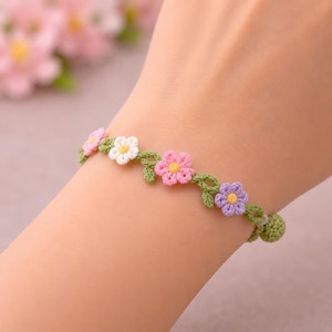 Könnte beinhalten: Ein gehäkeltes Armband mit floralem Design. Das Armband hat eine grüne Ranke mit rosa, weißen und lila Blumen mit gelben Zentren. Das Armband wird am Handgelenk getragen.