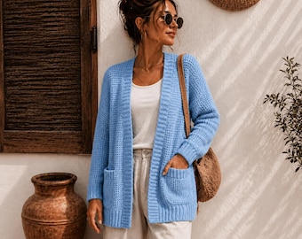 May include: A light blue open-front cardigan featuring a waffle-knit texture, long sleeves, and two front patch pockets. The garment is worn over a white top and light-colored trousers, paired with a round brown woven shoulder bag.