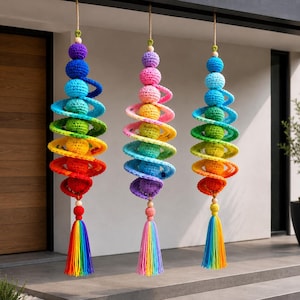 May include: Three hanging crochet wind spinners featuring a spiral design with stacked spheres in vibrant rainbow colors. Each ornament is suspended by a cord and finished with a multi-colored tassel at the bottom, showcasing a gradient of hues.