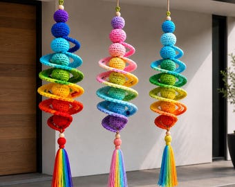 Puede incluir: Tres decoraciones colgantes con disposiciones verticales de esferas de ganchillo y elementos en espiral en degradados de colores arcoíris. Cada pieza está suspendida por un cordón y termina con una borla de hilo multicolor.
