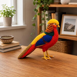May include: A vibrant, handmade crochet pheasant figurine. The bird features a yellow crest, a red body, and wings with yellow, blue, and black detailing. The pheasant is perched on a wooden surface, showcasing its intricate craftsmanship and colorful design.