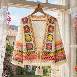 May include: A cream-colored crochet cardigan featuring a granny square pattern with multi-colored floral motifs in pink, yellow, and orange. The garment has long sleeves with horizontal stripes in pink, yellow, and green, and a front tie closure with tassels.