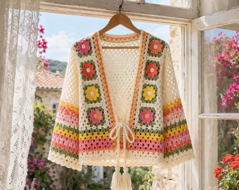 May include: A cream-colored crochet cardigan featuring a granny square pattern with multi-colored floral motifs in pink, yellow, and orange. The garment has long sleeves with horizontal stripes in pink, yellow, and green, and a front tie closure.