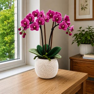 Könnte beinhalten: Eine handgefertigte Orchideenpflanze aus Häkelarbeit in einem weißen Topf. Die Orchidee hat leuchtend magenta Blüten, grüne Blätter und braune Stiele. Der Topf ist rund und strukturiert und steht auf einer Holzoberfläche.