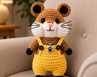 May include: A crocheted hamster plush toy with brown and white yarn, wearing bright yellow overalls with black buttons. The hamster has black eyes, a pink nose, black whiskers, and a silver chain necklace featuring a black paw print charm.