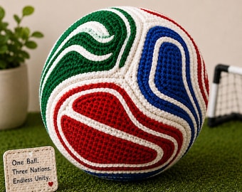 May include: A crocheted soccer ball featuring swirling patterns of red, white, blue, and green yarn. A small square tag sits nearby with the text: One Ball. Three Nations. Endless Unity. with a small red heart icon at the bottom.