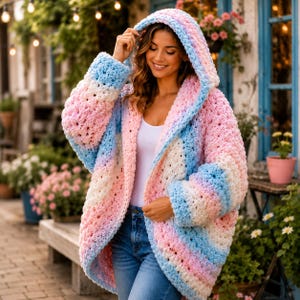 May include: A chunky, oversized hooded cardigan featuring a soft, variegated yarn pattern in pastel shades of pink, blue, and white. The knit garment has long sleeves, an open front design, and a large, cozy hood, worn over a white top and blue jeans.