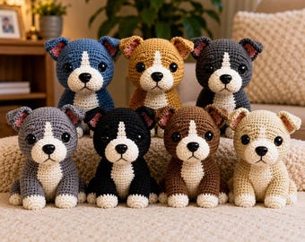 May include: A collection of seven crocheted puppy figurines in various colors including blue, tan, dark gray, light gray, black, brown, and cream. Each plush dog features a white chest, white muzzle, black button eyes, and pink inner ears.
