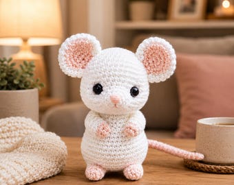 May include: A white crocheted mouse plush toy with large round ears featuring pink centers, black safety eyes, a small pink nose, and pink accents on the paws and feet. The mouse has a long pink crocheted tail extending to the right side.