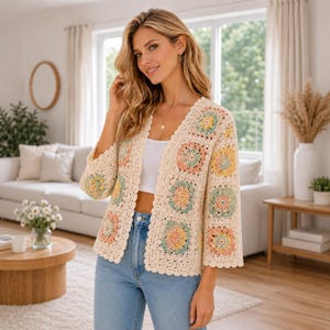 Könnte beinhalten: Cremefarbene Häkeljacke mit bunten quadratischen Mustern. Die offene Jacke hat einen gewellten Rand und Dreiviertelärmel. Die Quadrate haben Pastellfarben in Gelb, Rosa, Blau und Grün. Die Jacke wird mit einem weißen Top und blauen Jeans getragen.