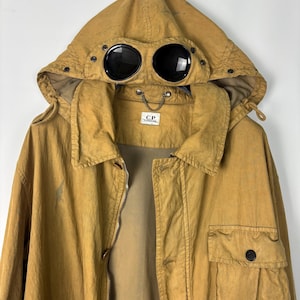 May include: A mustard yellow hooded jacket featuring integrated black circular goggles on the hood, a front zipper closure, a chest pocket with a button flap, and a C.P. Company brand label located at the inner neckline with a small metal chain detail.