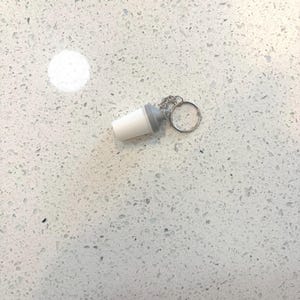 May include: A white and gray miniature shaker bottle keychain. The keychain features a small, detailed replica of a shaker bottle, with a white body and a gray top. The keychain is attached to a silver-colored keyring.
